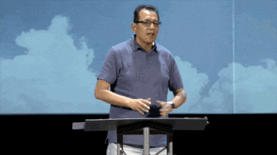 a man in a blue shirt stands at a podium with a microphone