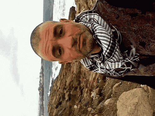 a man with a scarf around his neck is standing on a rocky cliff near the ocean