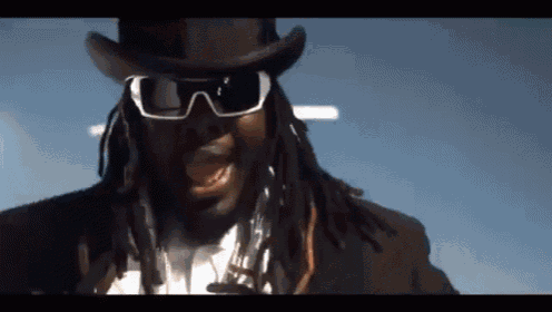 a man with dreadlocks wearing a top hat and sunglasses looks at the camera