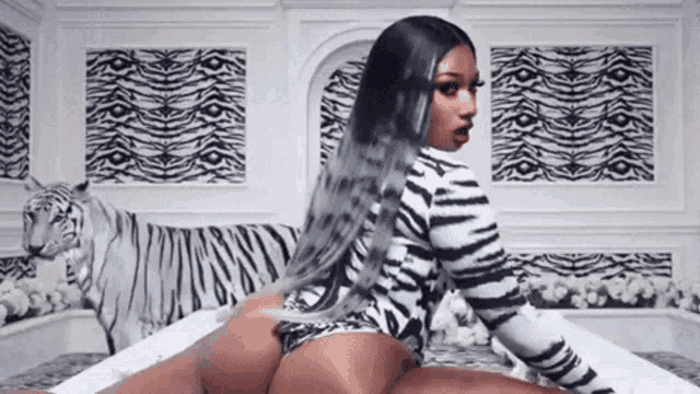 a woman in a zebra print bodysuit is sitting on a bed with a white tiger behind her .