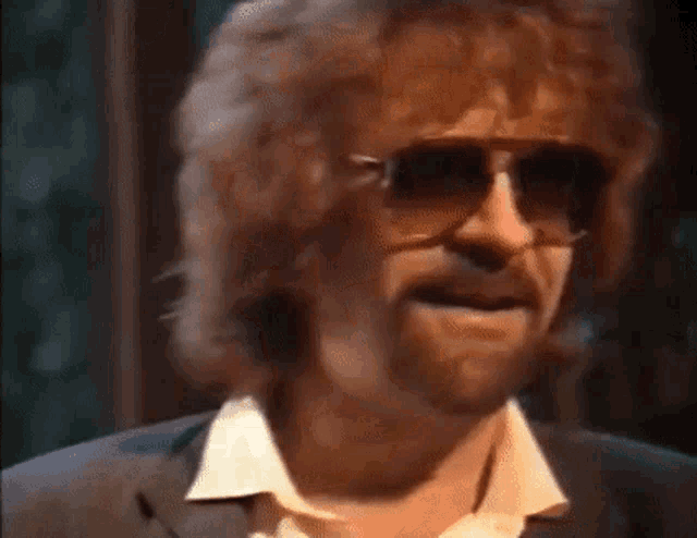 a man with a beard and sunglasses is wearing a suit and a white shirt .
