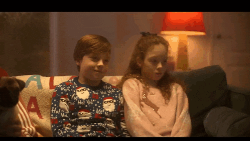 a boy and a girl are sitting on a couch wearing christmas sweaters that say ho ho ho