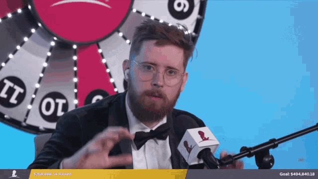 a man in a tuxedo is talking into a microphone in front of a spinning wheel with the number 6 on it