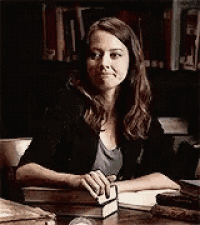 a woman is sitting at a desk in front of a bookshelf holding a book .