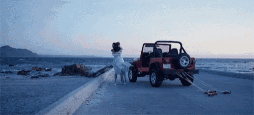 a woman in a white dress stands next to a red jeep