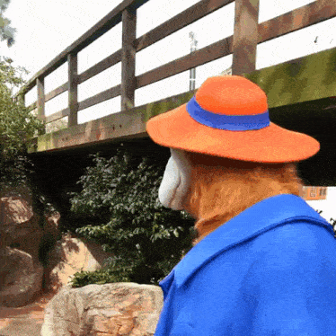 a teddy bear wearing an orange hat and a blue ribbon stands in front of a bridge