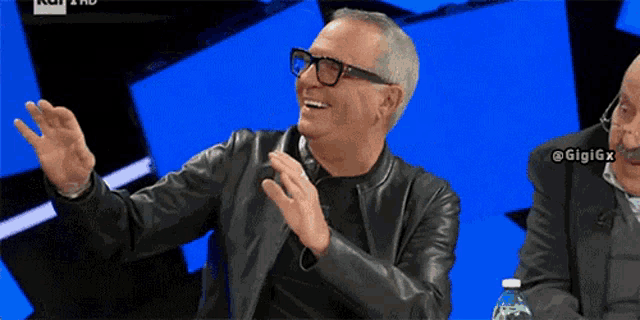 a man wearing glasses and a leather jacket laughs while sitting next to another man with a bottle of water