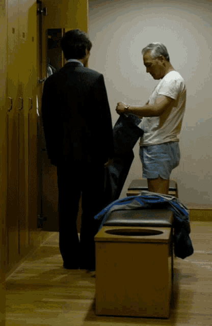 a man in a suit and a man in a white shirt are in a dressing room