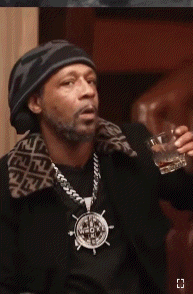 a man wearing a hat and a necklace with a steering wheel pendant is drinking a glass of whiskey