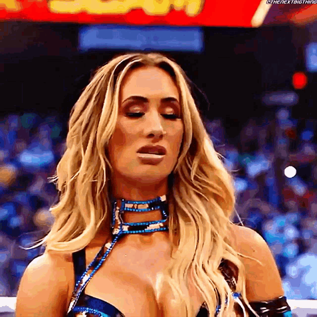 a woman with blonde hair and a blue choker stands in a wrestling ring with her eyes closed