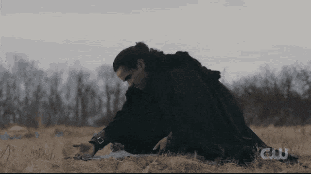 a man in a black cape is kneeling down in a field with a cw logo in the background