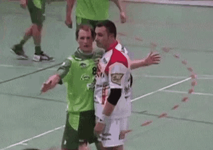 a man in a green shirt is jumping in the air while a man in a white shirt is sitting on the floor .