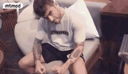 a man with a tattoo on his arm is sitting on a couch with his legs crossed .