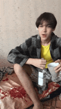 a young man sits on a bed holding a cup and a bottle of vodka