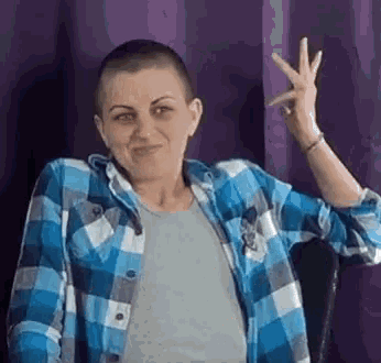 a woman with a shaved head is wearing a blue and white plaid shirt and making a peace sign .