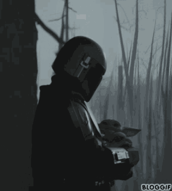 a man in a helmet is holding a baby in a dark forest with the words bloggif on the bottom right