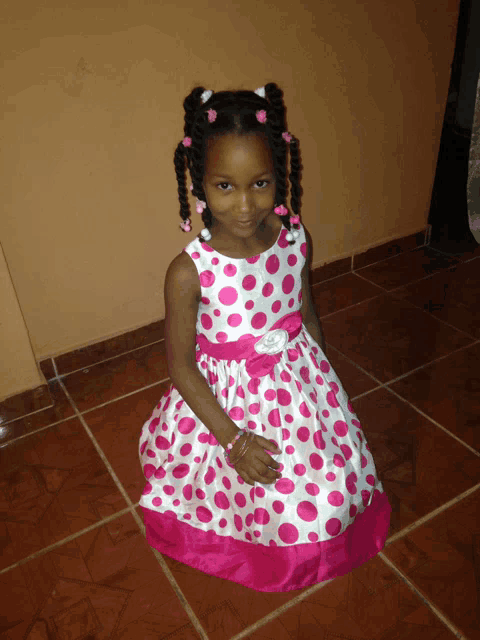 a little girl wearing a pink and white polka dot dress sits on a tiled floor
