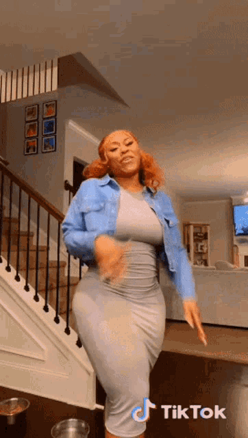 a woman in a grey dress is dancing in a living room with stairs in the background and a tiktok sticker on the bottom