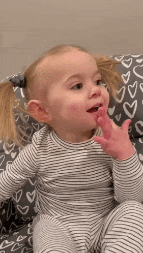 a little girl in a striped shirt is sitting on a couch with her hand to her mouth .