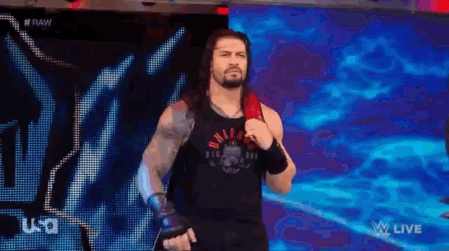 roman reigns is wearing a black tank top and holding a red belt while walking on a stage .