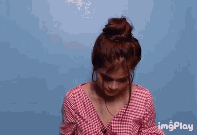 a woman in a red plaid shirt is looking down with a blue background and the word imgplay behind her