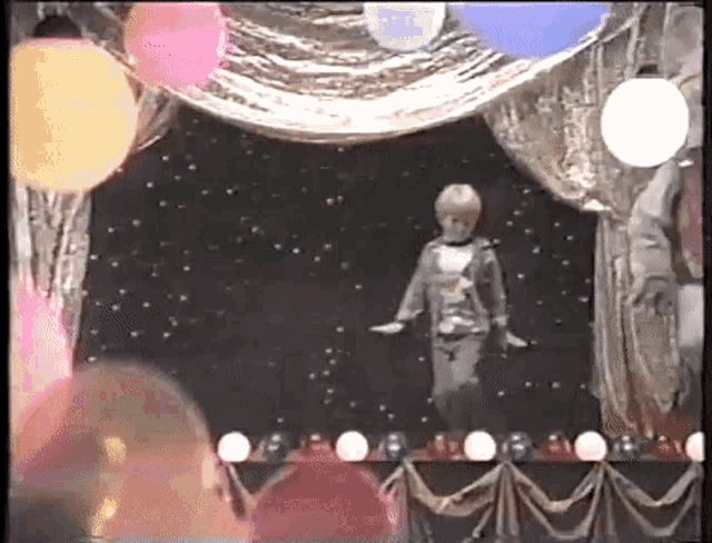 a little girl is standing on a stage with balloons and lights .