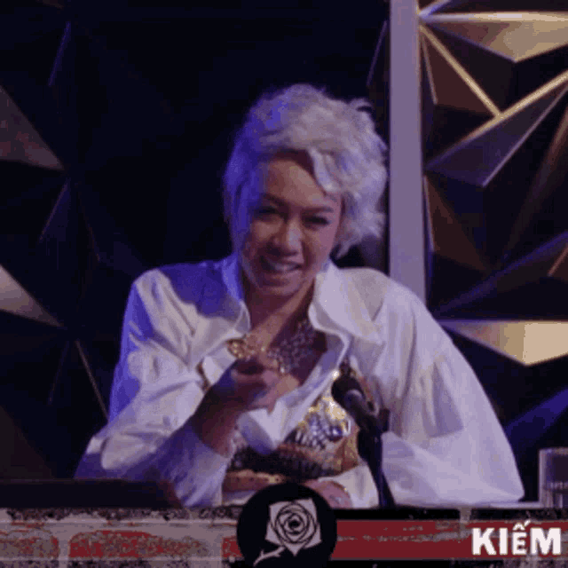 a woman in a white shirt is sitting in front of a microphone and a sign that says kiem