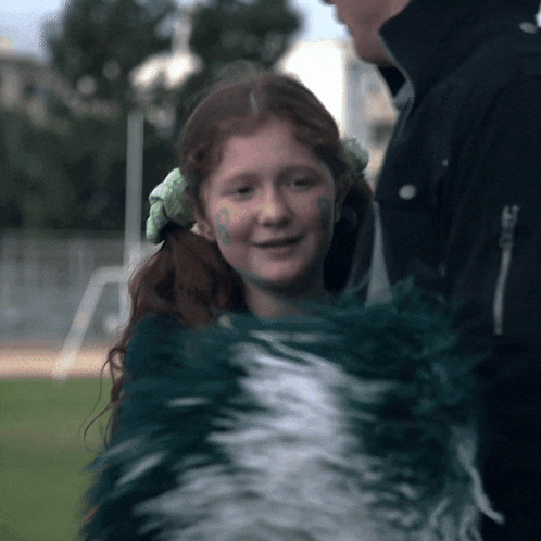 a girl with green paint on her face holds a green and white cheerleading pom pom