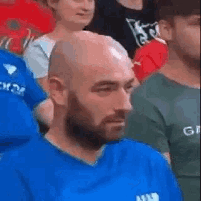 a bald man with a beard is sitting in a crowd of people wearing a blue shirt .