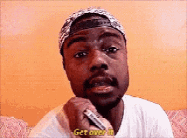 a man wearing a hat and a bandana is talking into a microphone and saying get over it .