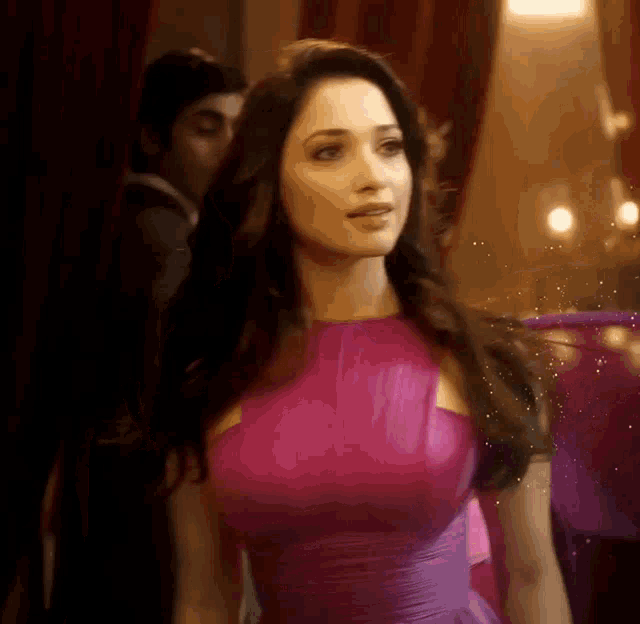 a woman in a purple dress is standing in a room with a man behind her .