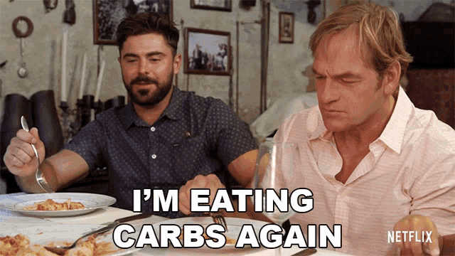two men are sitting at a table with plates of food and a caption that says " i 'm eating carbs again "