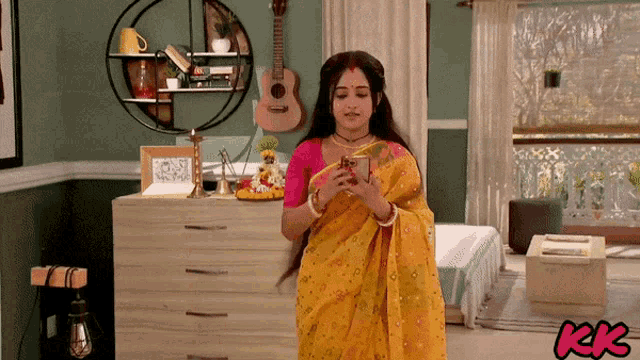 a woman in a yellow sari is looking at her phone in a room with kk written on the bottom