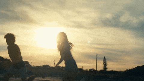a boy and a girl are running towards the sun at sunset