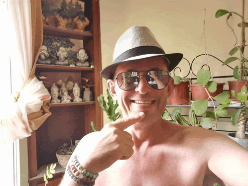 a shirtless man wearing a hat and sunglasses is pointing at his face