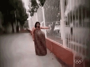 a woman in a long dress is standing next to a fence with the olympic rings in the background .