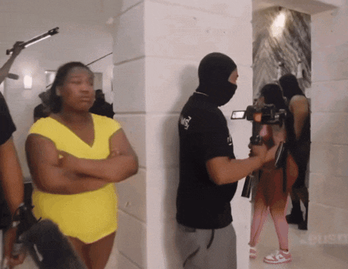 a man wearing a mask holds a camera while a woman in a yellow dress stands behind him