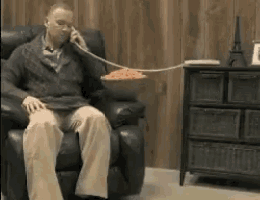 a man is sitting in a chair talking on a phone with a bowl of spaghetti attached to his mouth .