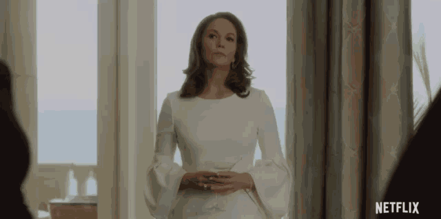 the woman is wearing a white dress and standing in front of a window .