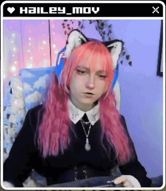 a girl with pink hair and cat ears is sitting in a chair and looking at the camera .