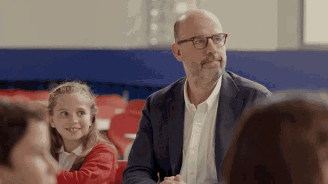 a man wearing glasses sits in a classroom with a girl