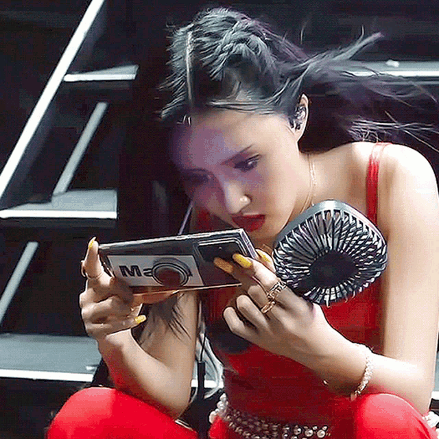 a woman in a red dress is holding a fan and looking at a cell phone with the letter m on the screen