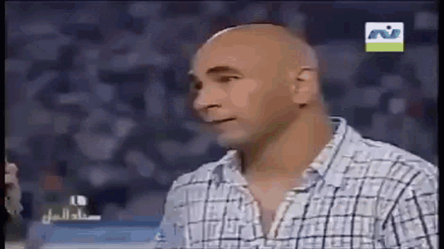 a bald man wearing a plaid shirt is talking to someone on a television screen .