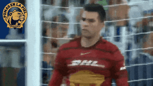 a man in a red pumas en la piel jersey stands in front of a goal