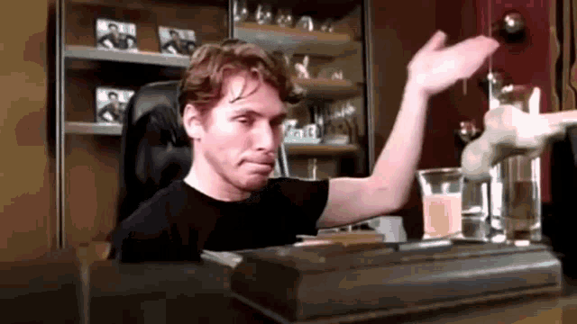a man in a black shirt is sitting at a table with his hand up .