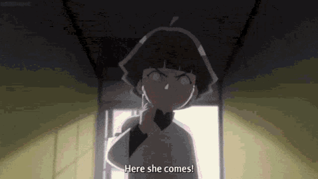 a girl in a white shirt is standing in a hallway and says `` here she comes '' .