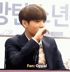 a young man in a suit is sitting at a table with his hand on his mouth and says fan oppa .