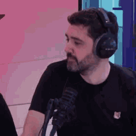 a man with a beard wearing headphones and a black shirt is sitting in front of a microphone .