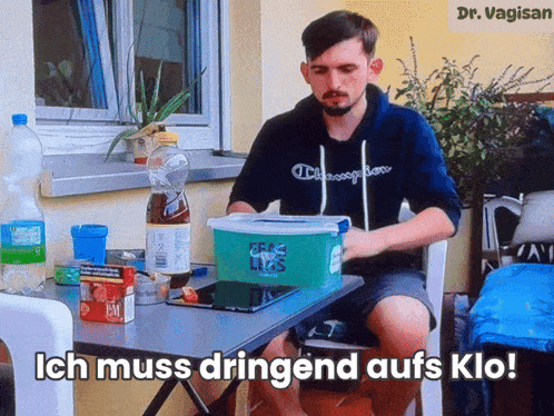 a man sits at a table with a box on it that says ich muss dringend aufs klo on it