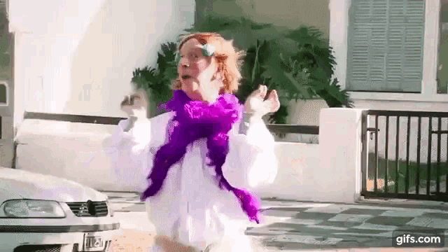 a woman wearing a white shirt and a purple boa is dancing in front of a car .
